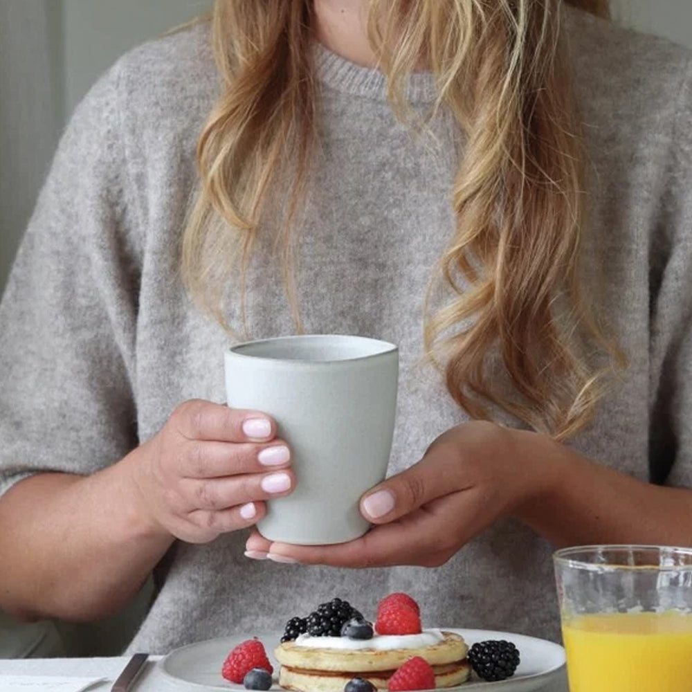 Vrouw met grijze trui houdt mok vast met ontbijt op tafel 