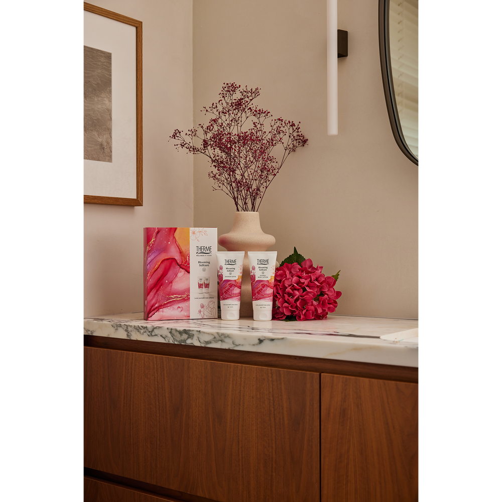 Therme Blooming Selfcare geschenkset met douchegel en bodylotion op een marmeren badkamermeubel, naast roze bloemen en een vaas met droogbloemen. Een sfeervol verzorgingscadeau voor een verwenmoment thuis.