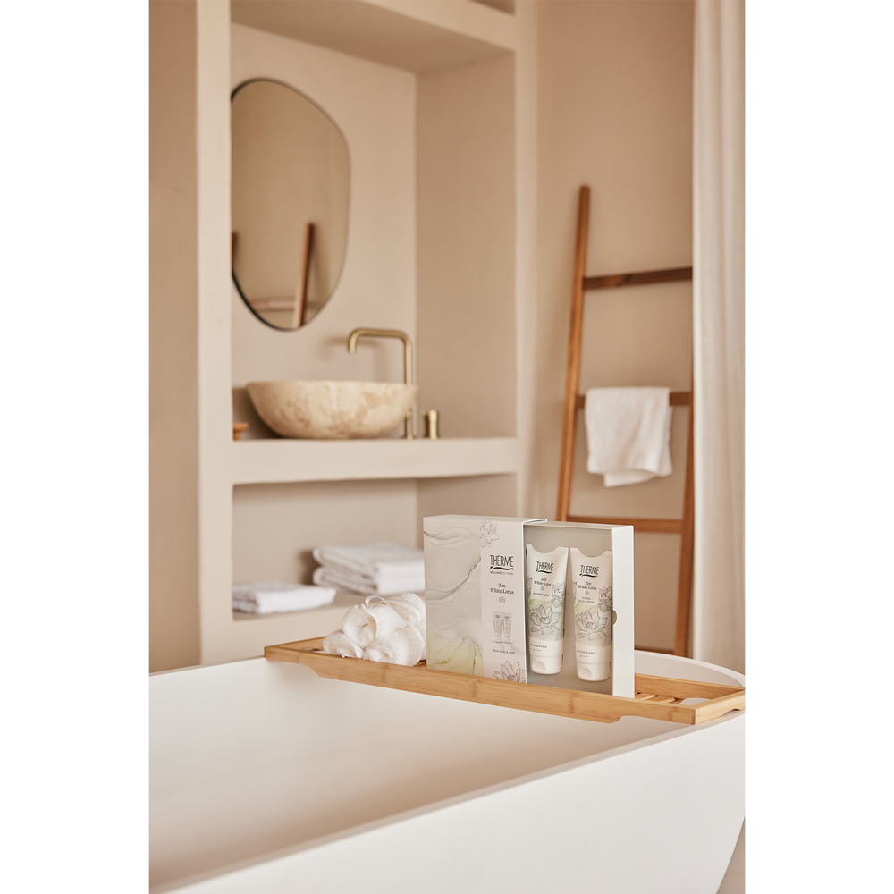 Therme Zen White Lotus geschenkset met douchegel en bodylotion op een houten badplank, naast een opgerolde witte handdoek. Een sfeervol wellnessmoment in een lichte badkamer met vrijstaand bad.