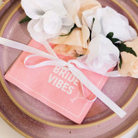 een roze bordje met een servet met bride vibes erop en een lint met bloemen erbij.